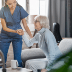 The shot is of a patient sitting up in a home care setting. It focuses on the caretaker lifting up the patient.
