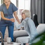 The shot is of a patient sitting up in a home care setting. It focuses on the caretaker lifting up the patient.