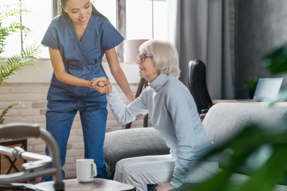 The shot is of a patient sitting up in a home care setting. It focuses on the caretaker lifting up the patient.