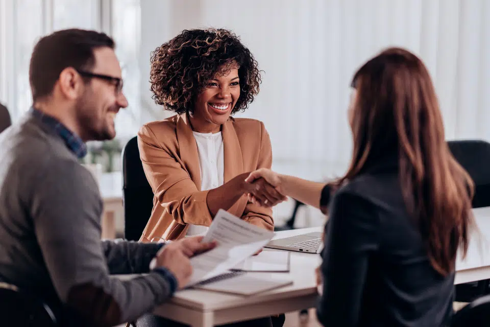 A woman sitting across from 2 staff members at a table. All are wearing business clothing and meeting each other