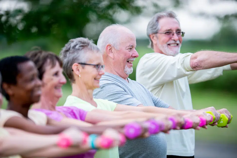 Group of smiling older adults exercising outdoors with small hand weights.