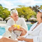 Smiling senior couple in white robes clinking glasses of orange juice while relaxing on lounge chairs by a pool.