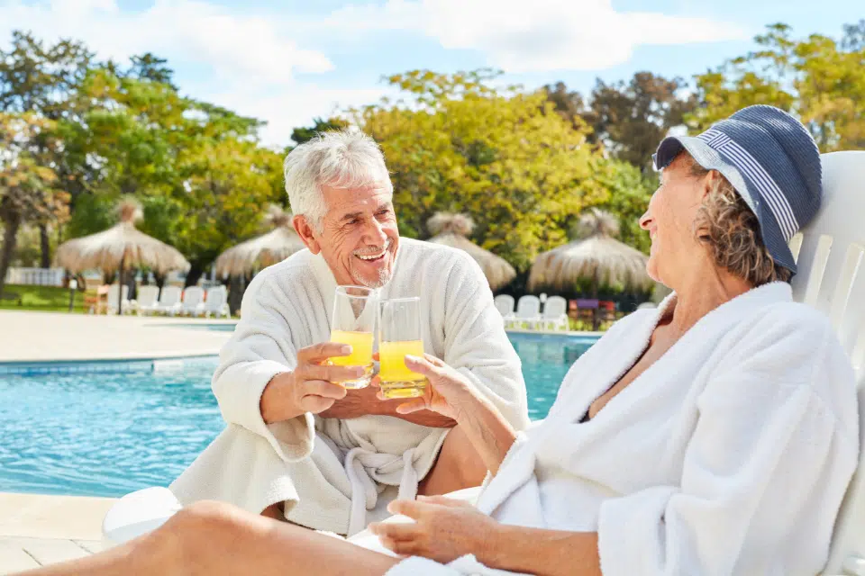 Smiling senior couple in white robes clinking glasses of orange juice while relaxing on lounge chairs by a pool.